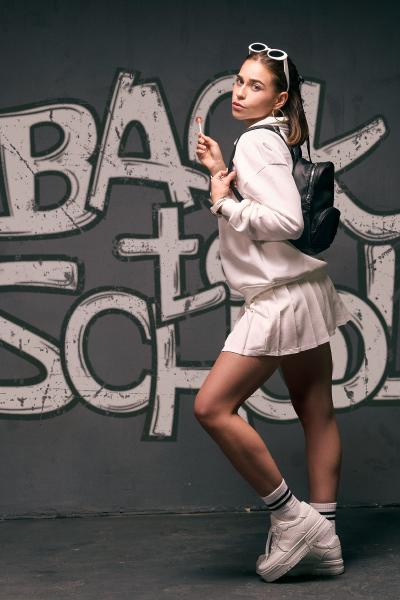Kobieta w białej bluzie i plisowanej spódniczce tenisowej z lizakiem w dłoni, plecak, napis Back to School na murze – matee.photo