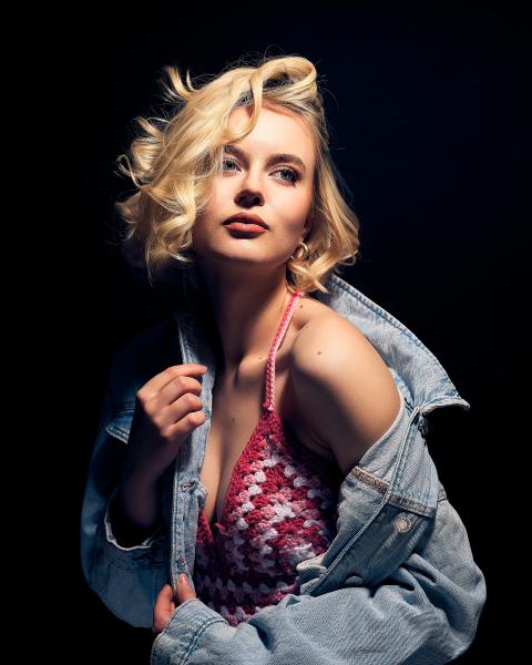 Kobieta w rozpiętej kurtce jeansowej odsłaniającej różowo-biały szydełkowy top, falowany blond bob na czarnym tle – matee.photo