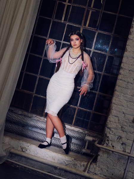Kobieta w stylizacji lalki z tiulowym rękawem i perłowymi naszyjnikami stoi przy przemysłowym oknie z warkoczami – matee.photo