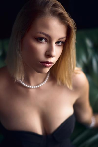 Portret blond kobiety w czarnym gorsecie i sznurze pereł na tle ciemnozielonej pikowanej kanapy, miękkie światło – matee.photo