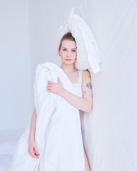 Kobieta owinięta białą kołdrą z poduszką przy głowie opiera się o białą ścianę, tatuaże na ramieniu – matee.photo