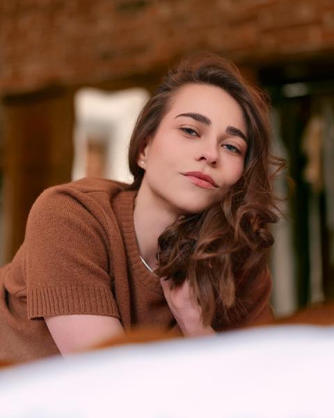Portret kobiety w brązowym swetrze opartej na dłoni, kasztanowe fale, ciepłe rozmyte wnętrze w tle – matee.photo