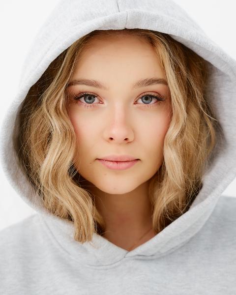Portret kobiety w szarym kapturze, blond fale do ramion, niebieskie oczy, naturalny makijaż na jasnym tle – matee.photo