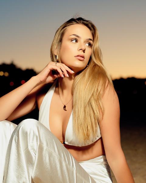 Blond kobieta w białym topie z wiązaniem na szyi siedzi na plaży o zmierzchu, dłoń przy twarzy, światła w tle, plaża w Gdańsku-Brzeźnie – matee.photo