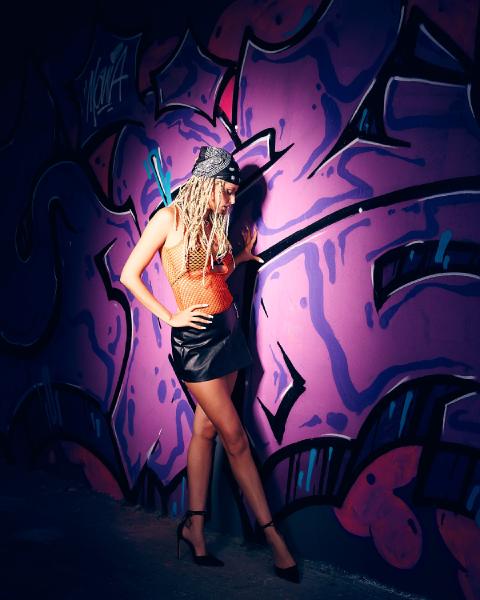 Fioletowe graffiti nocą w świetle flesza, kobieta z warkoczykami w bandanie, pomarańczowym topie i skórzanej spódniczce opiera się o ścianę, Warszawa – matee.photo