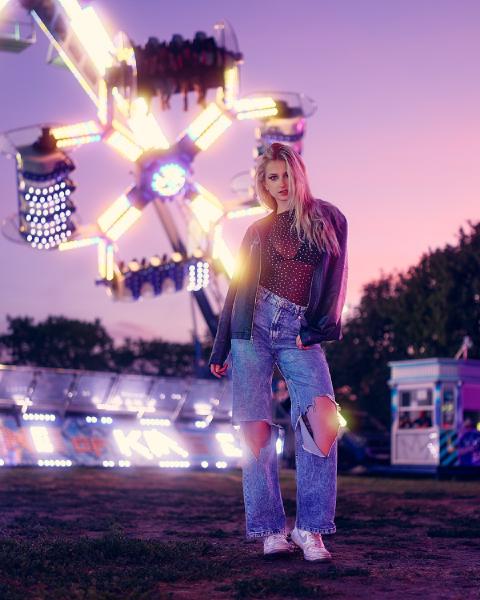 Blond kobieta w skórzanej kurtce, siateczkowym topie i podartych jeansach stoi przed świecącą karuzelą w wesołym miasteczku o zmierzchu, Warszawa – matee.photo