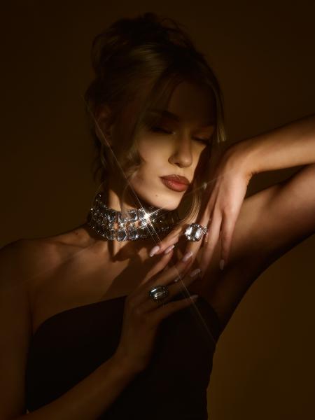 Modelka z upiętymi włosami dotyka chokera z kryształów, dłoń z pierścieniem uniesiona nad głową, ciepłe światło – matee.photo