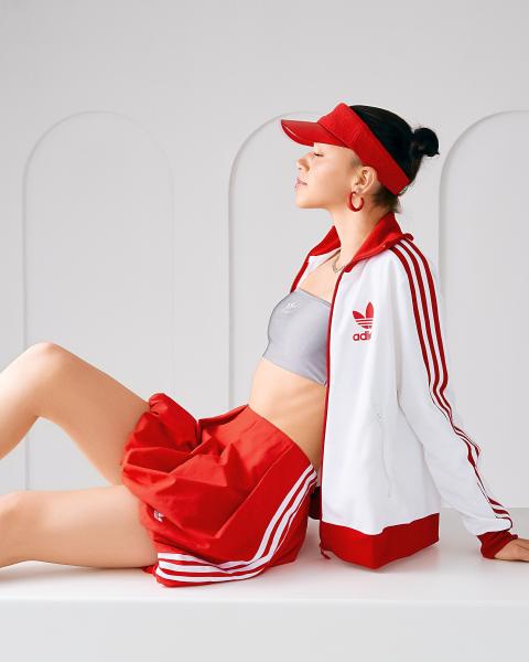 Modelka w czerwonym daszku adidas i bluzie z lampasami siedzi z zamkniętymi oczami, czerwone szorty i szary top – matee.photo