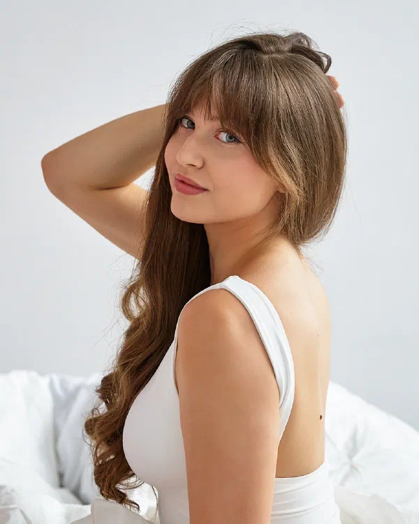 Biała pościel i miękkie światło, portret kobiety z grzywką w białym body z dłonią we włosach – matee.photo