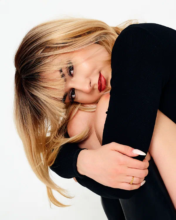 Białe tło, blond kobieta w czarnym swetrze opiera głowę o kolano, czerwone usta – matee.photo