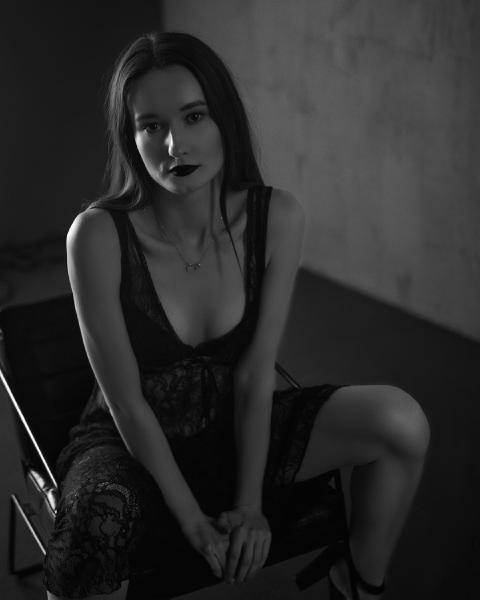 Czarno-biały portret przy ciemnej ścianie, kobieta w koronkowej sukience siedzi pochylona – matee.photo