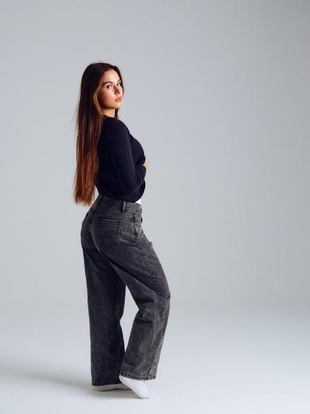 Modelka odwrócona tyłem w czarnym swetrze i szerokich szarych jeansach, spojrzenie przez ramię – matee.photo