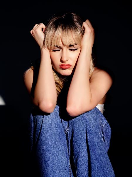 Blond modelka w jeansowej sukience siedzi w ciemnej scenerii, ręce przy twarzy, dramatyczne światło – matee.photo