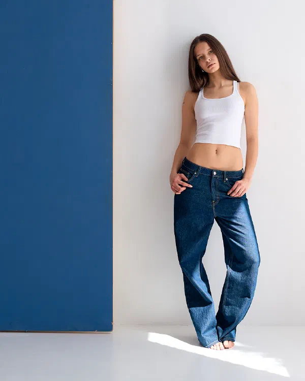 Modelka w białym crop topie i ciemnych jeansach oparta o białą ścianę obok granatowej planszy, dłonie w kieszeniach – matee.photo
