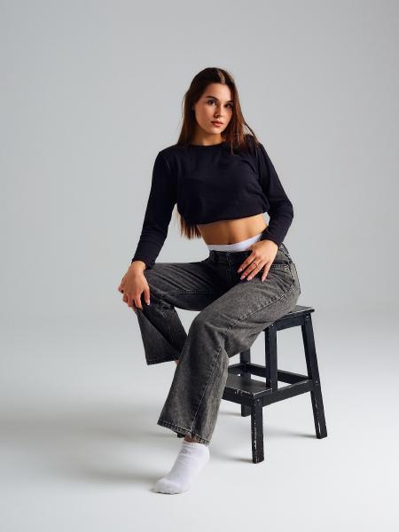 Modelka siedzi na czarnym stołku w krótkiej bluzce i szarych jeansach, noga oparta na kolanie – matee.photo